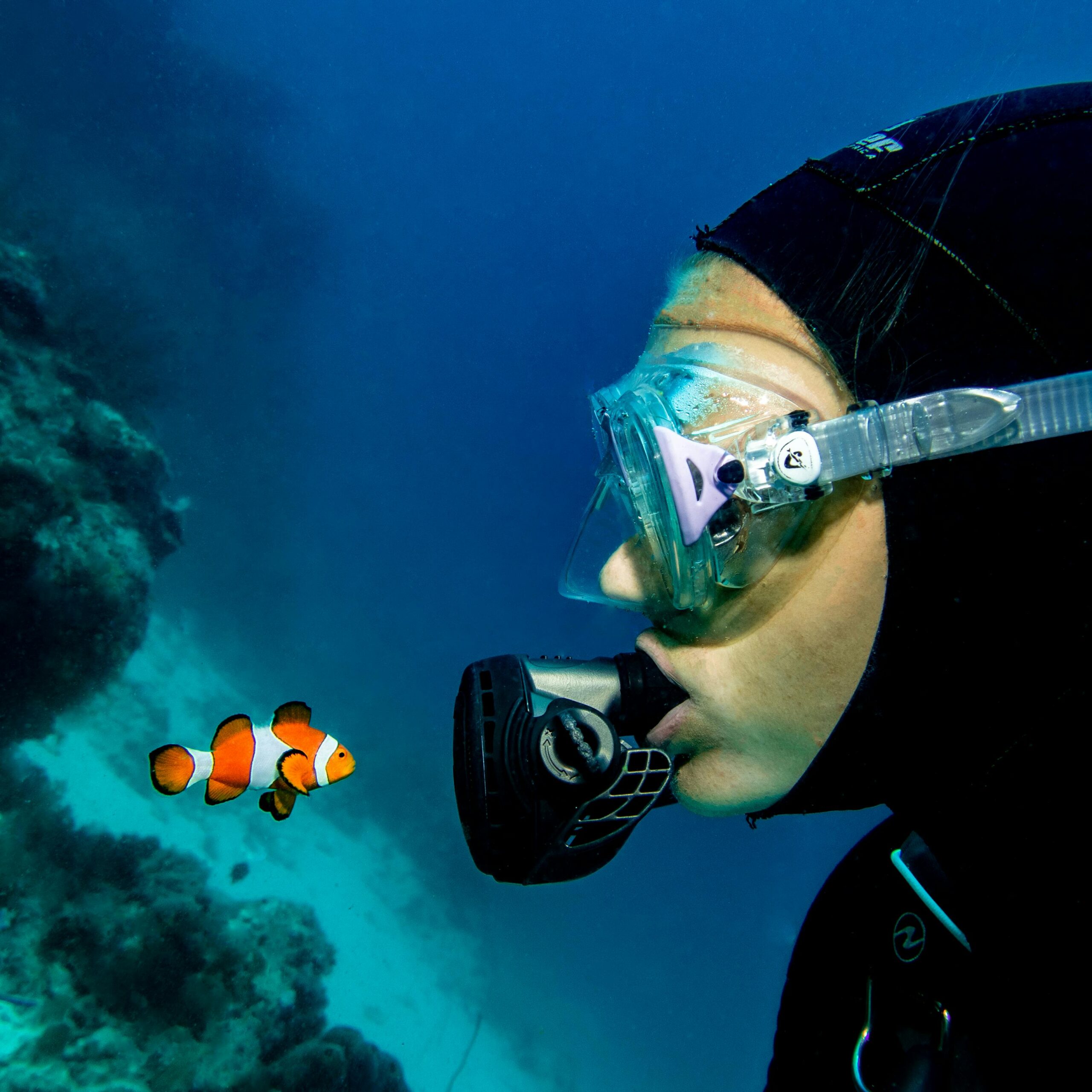 A scuba diver wearing goggles closely observes a clownfish in the ocean.
